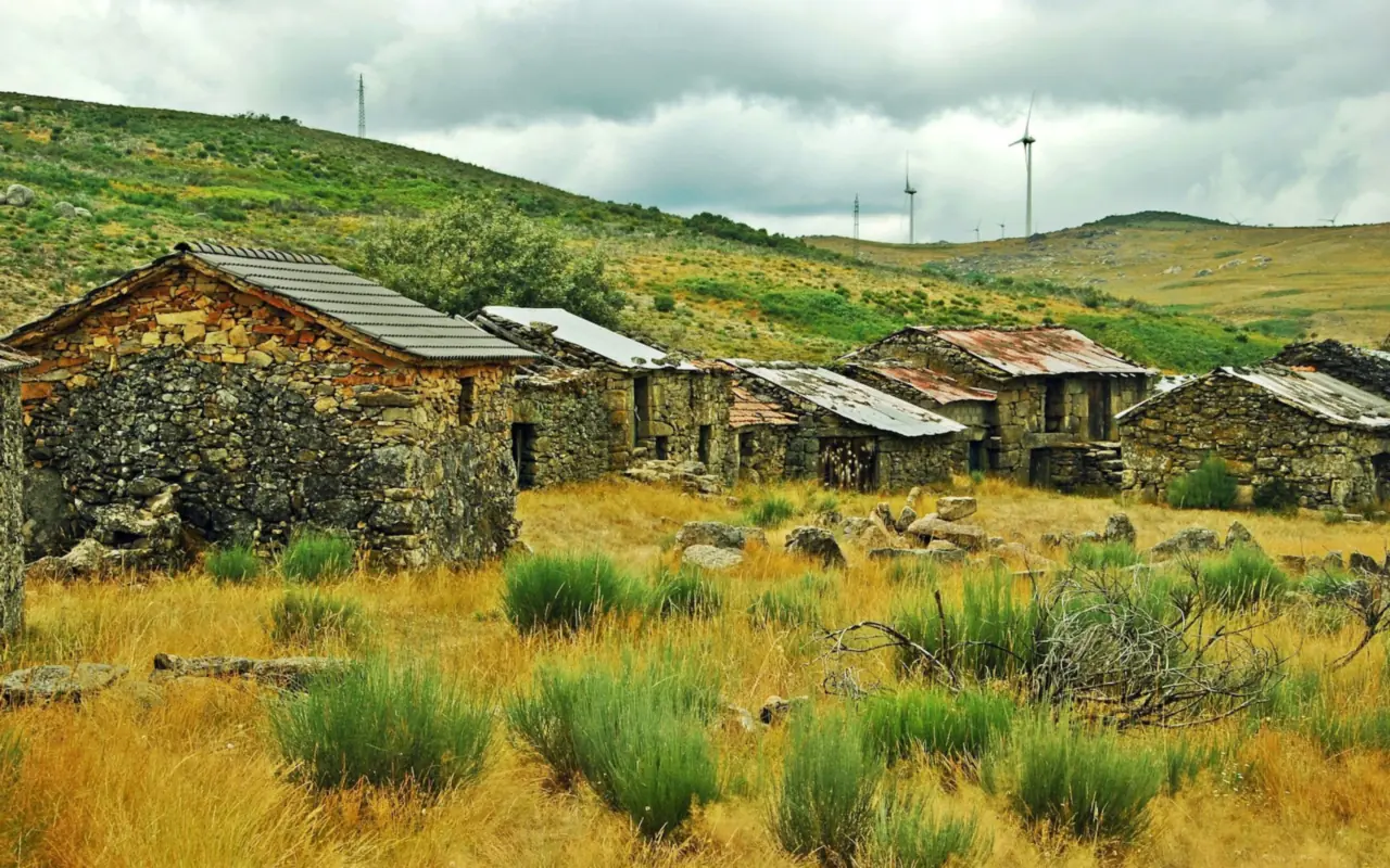 A cluster of abandoned dry-stone houses with tin and slate roofs on a grassy hillside in rural Portugal