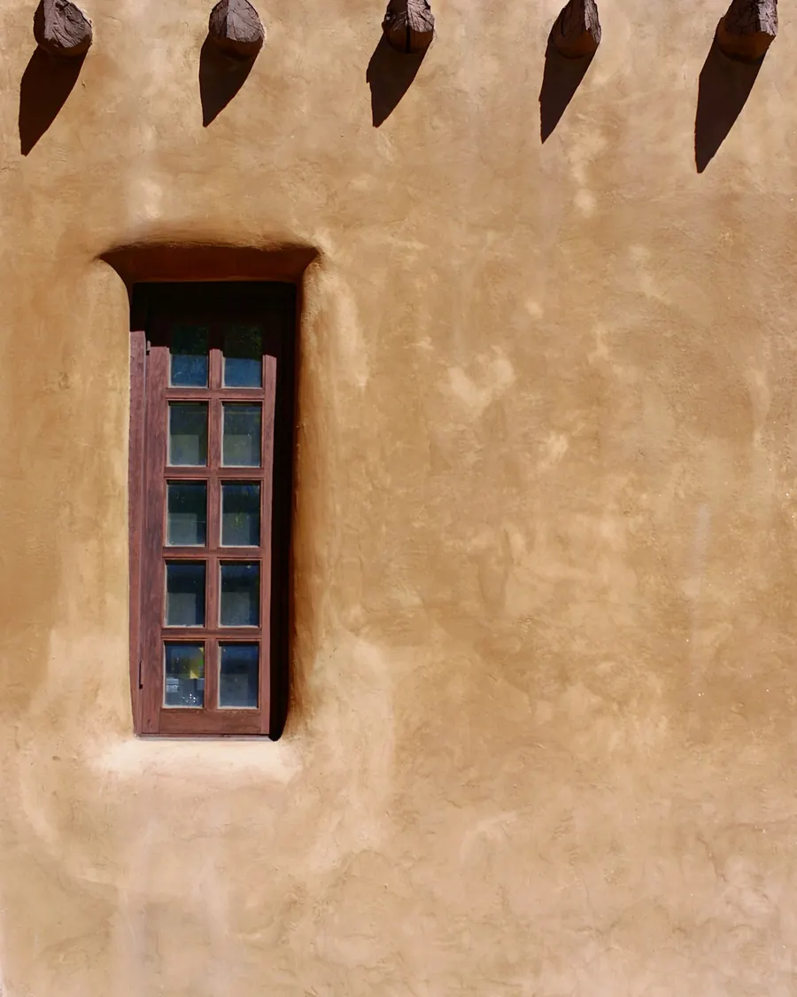 A smooth, hand-finished earth-toned render wall with a narrow deep-set timber window and projecting roof beams